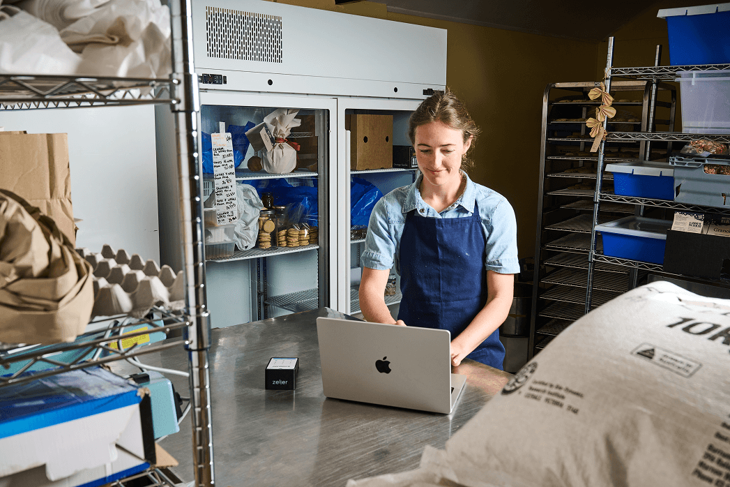 A person in an apron operates a laptop in an organised kitchen, surrounded by supplies and shelves near a refrigerator.