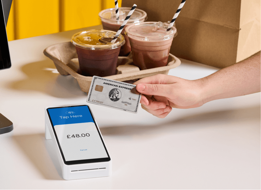 Hand holding an American Express card near a payment terminal showing £48.00, with takeaway drinks in the background.