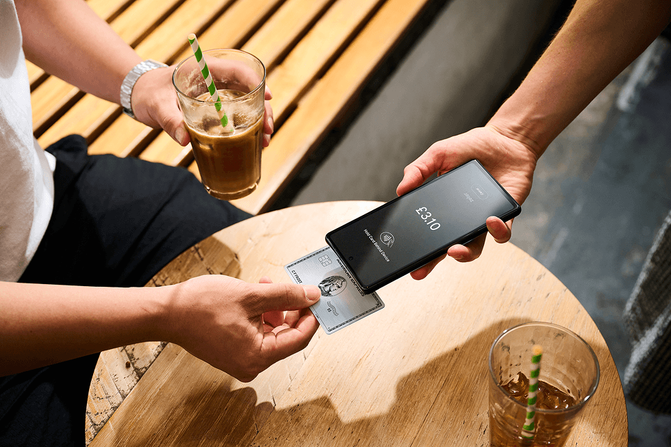 Person pays £3.10 with credit card using smartphone on table; holding iced coffee with striped straw.