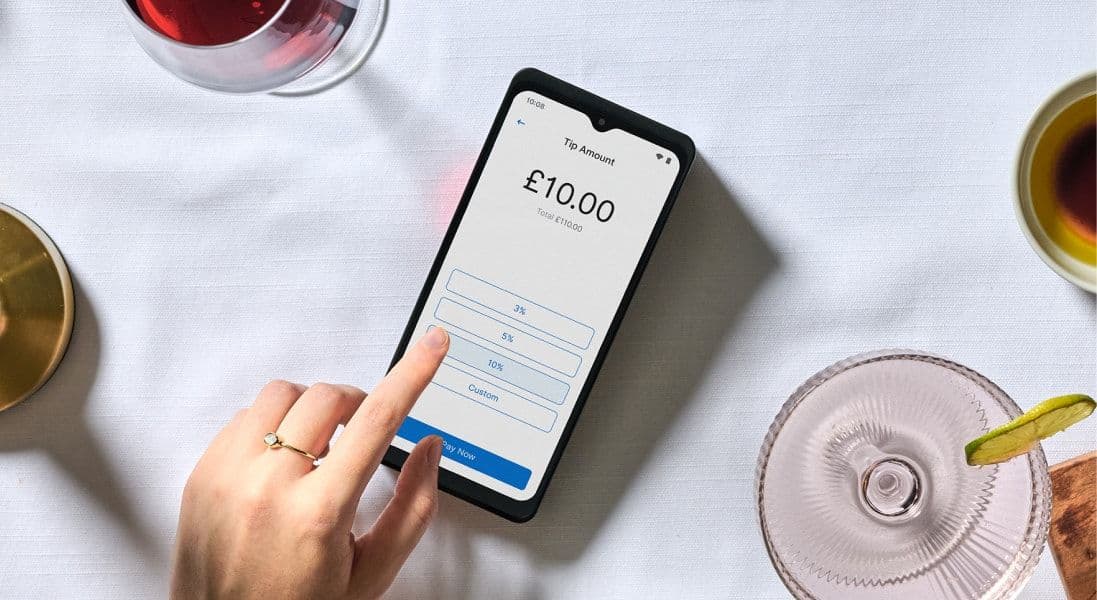 A person selects a tip amount on a £10 bill via a smartphone app, surrounded by wine, tea, and a cocktail on a white tablecloth.